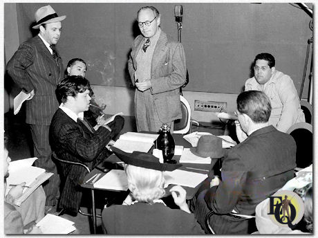 Orson Welles zit aan een tafel en rookt zijn pijp, met zijn ster, komiek Jack Benny, staand in het midden, en anderen tijdens de repetitie van June Moon in het radiostation KNX voor de uitzending van de CBS Campbell Playhouse, Hollywood, Californië, 24 maart 1940.