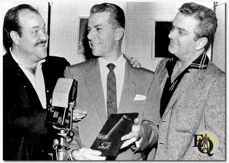 "Gunsmoke" is een Amerikaanse westernserie voor radio en televisie, gecreëerd door regisseur Norman Macdonnell (R) en schrijver John Meston. De verhalen spelen zich af in en rond Dodge City, Kansas, tijdens de kolonisatie van het Amerikaanse Westen. Het centrale personage is de wetsdienaar Marshal Matt Dillon, vertolkt door William Conrad (L) op de radio en James Arness op televisie. Rex Koury (midden) componeerde het themalied. Hier is hij te zien terwijl hij de “Bud Shank Flute”-prijs ontvangt van "DownBeat Magazine" (2 feb 1957).