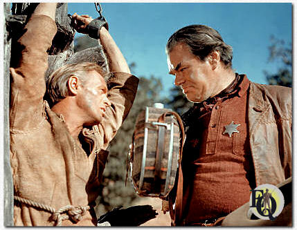 Overlevenden van een Apache-aanval op een woonwagen vertrouwen de gevangene van de sheriff, verkenner Comanche Todd (Richard Widmark), toe. Mathews is Sheriff Bull Harper in "The Last Wagon" (20th Century-Fox, 21 sep 1956).